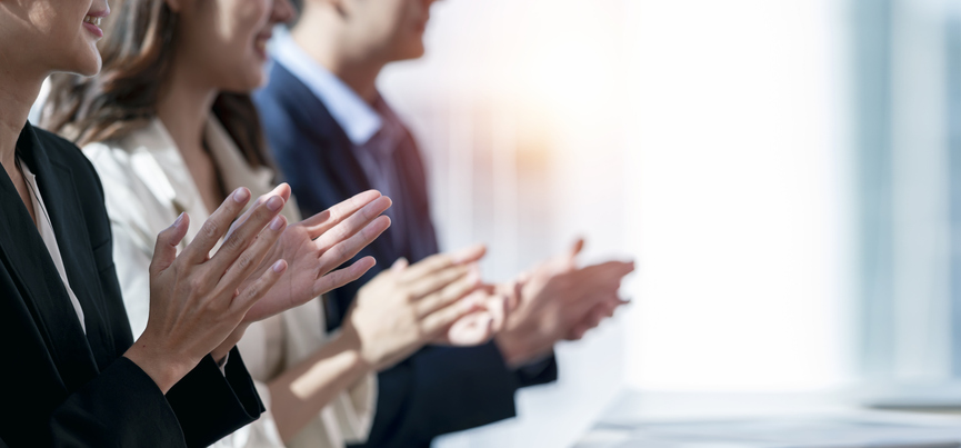Three people dressed in business attire are standing in a line while smiling and clapping. 
