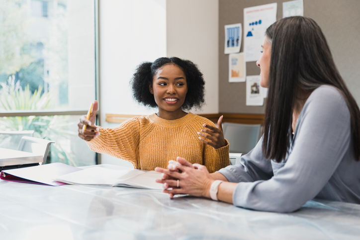 Two women are sitting at a table. While looking at each other, one has her arms open as if too demonstrate distance from one point to another.