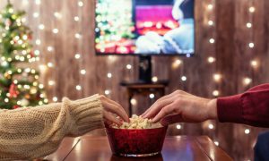 A close up photo of two people reaching into a bowl of popcorn that is sitting on a table. In the background, there is a Christmas tree, a TV, and strings of lights.
